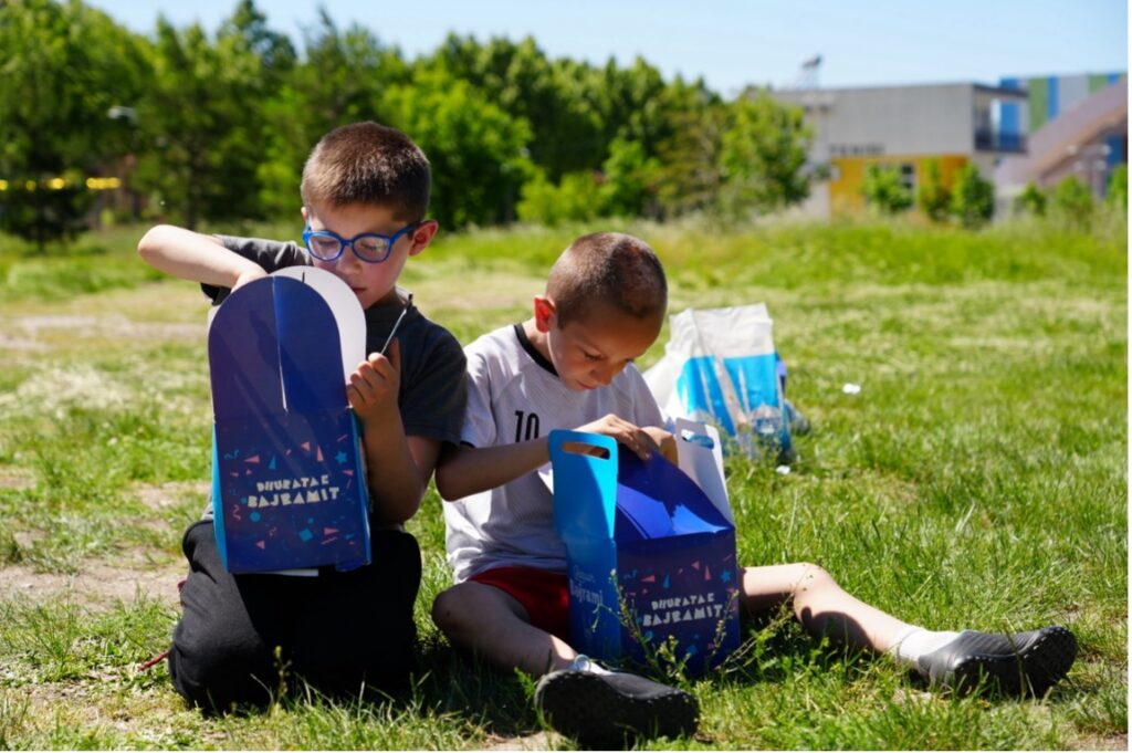 Due bambini aprono i loro pacchi regalo per l’Eid sull’erba in Albania. Fotografia di Hiba Ben Abdennabi.